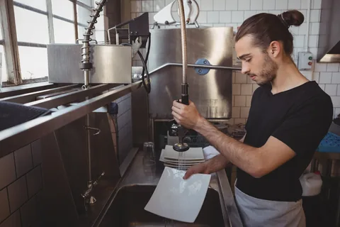 Young waiter washing plates while standing in cafe